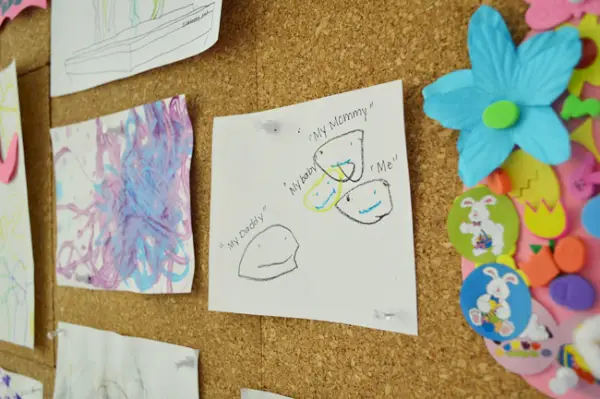 vaikų piešinys, kuriame mamytė tėtis aš ir kūdikis prisegtas prie milžiniškos kamštienos lentos sienos