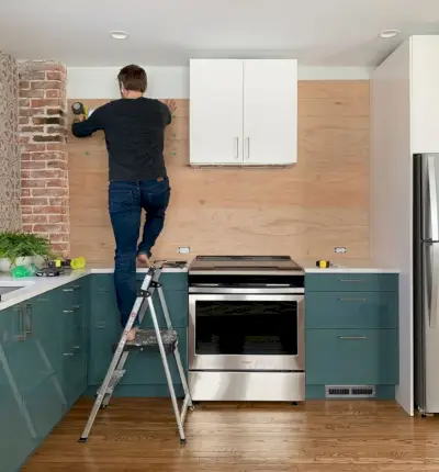 John staat op de ladder en spijkert de achterwand met houten panelen in de blauwe keuken