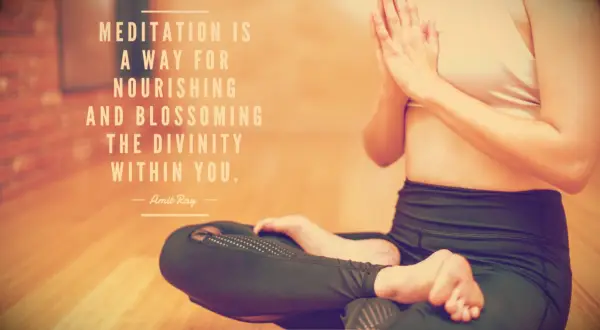 A woman in a yoga studio sitting in lotus pose with crossed legs and arms held in prayer 
